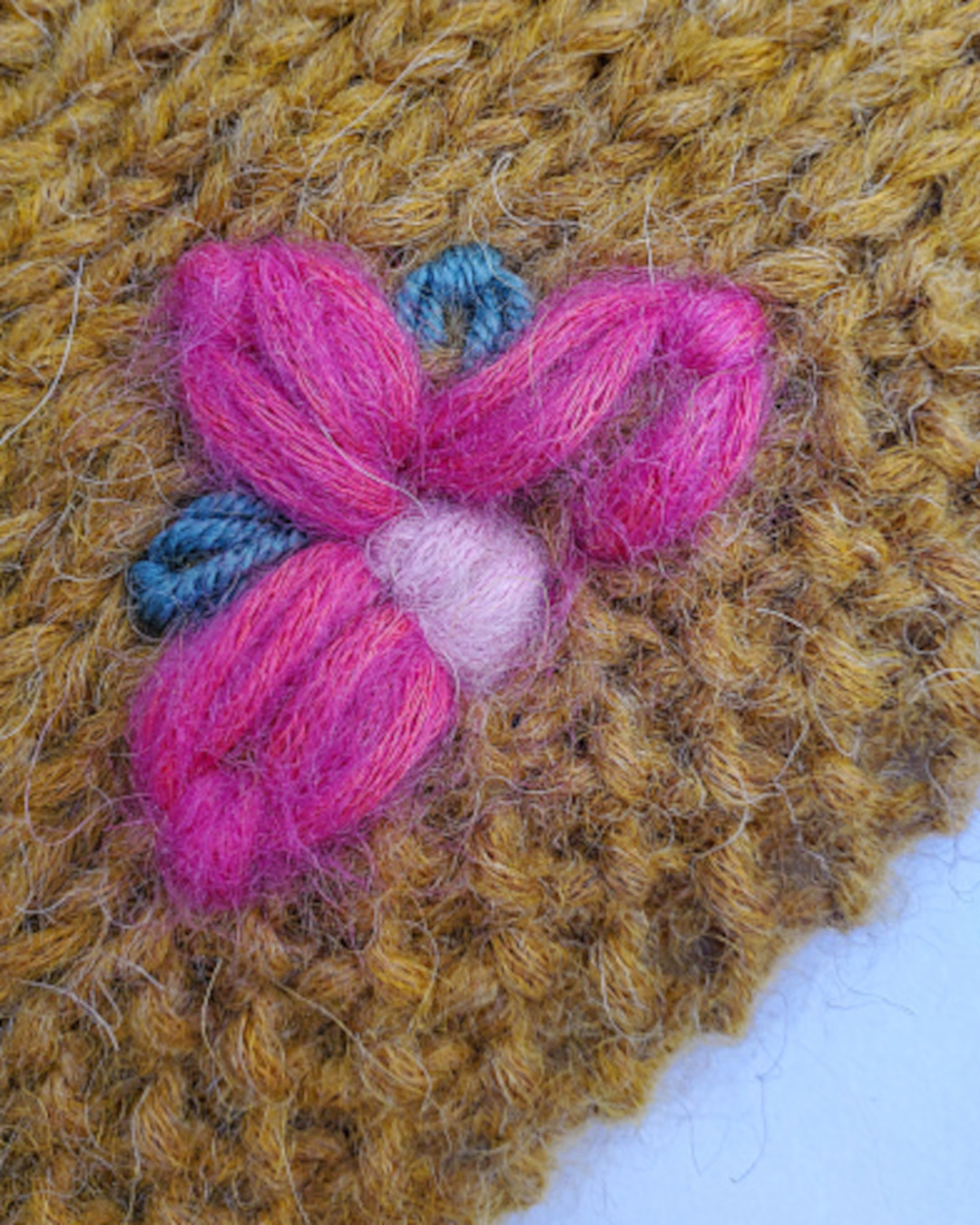 Detailansicht einer halben, pinken Blüte am Rand eines senfgelben Strickstückes