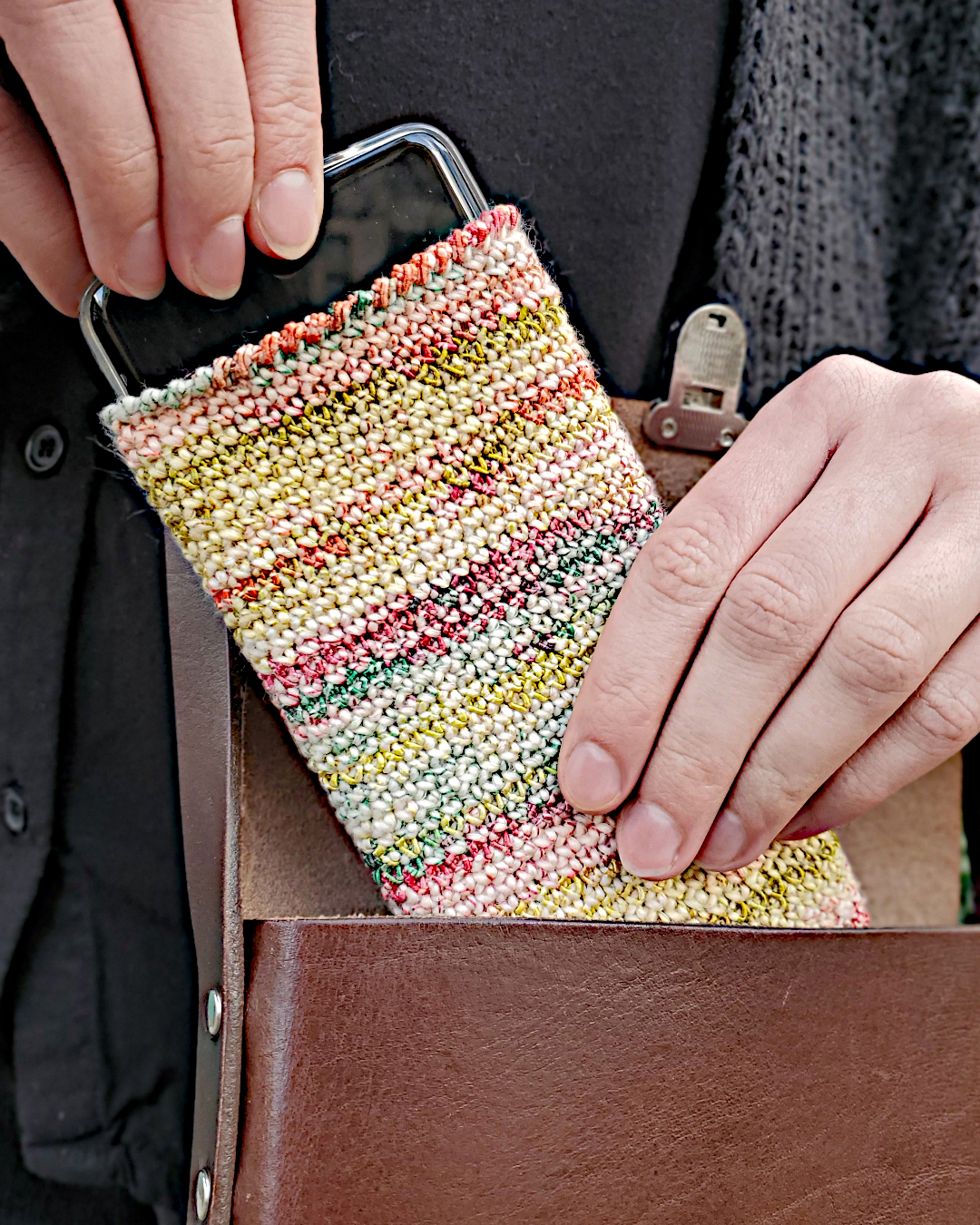 Ein Handy mit mehrfarbiger, gehäkelter Hülle wird aus einer Tasche genommen
