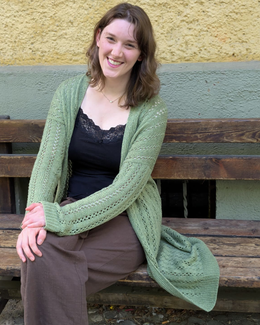 Modell sitzt auf einer Holzbank und trägt eine lange grüne Strickjacke mit vertikalem Lochmuster.