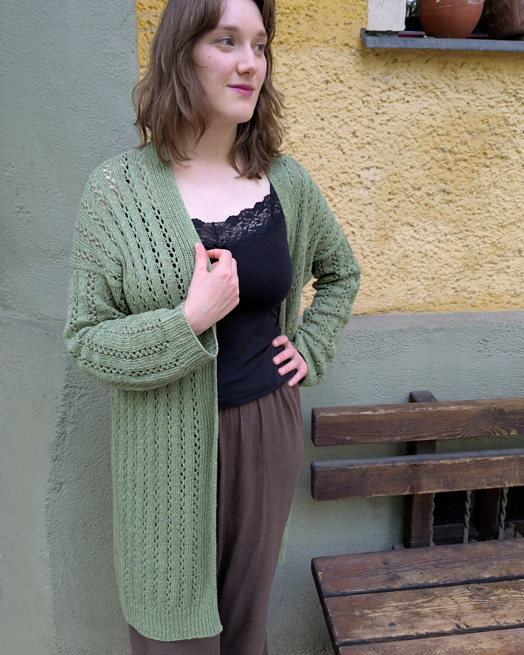 Modell steht neben einer Holzbank und trägt eine lange, grüne Strickjacke mit vertikalem Lochmuster